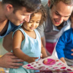 Two men and two young children engaging happily with a picture book. one man is pointing at the book, explaining something to the attentive children. they are seated indoors with natural light.