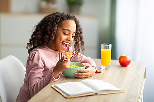 A joyful young girl eating cereal at a kitchen table, with an open book, an apple, and a glass of orange juice beside her. she is wearing a pink shirt and laughing while holding her spoon.