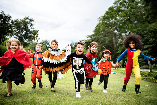 A group of joyful children in colorful costumes, including a superhero, astronaut, and skeleton, run playfully across a grassy park.