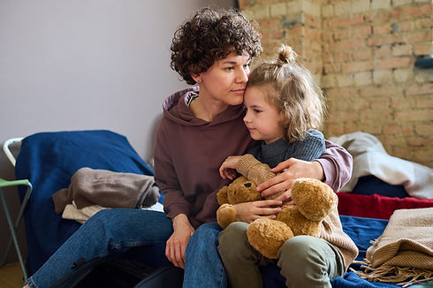 A woman with curly hair and a child holding a teddy bear sit closely on a sofa, sharing a tender moment in a cozy, warmly lit room with a brick wall background.