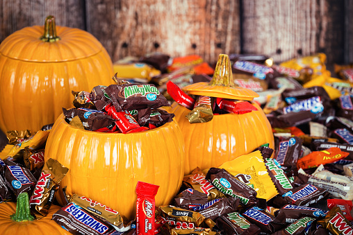 Two ceramic pumpkins overflowing with various halloween candies like snickers, milky way, and m&ms on a rustic wooden backdrop.