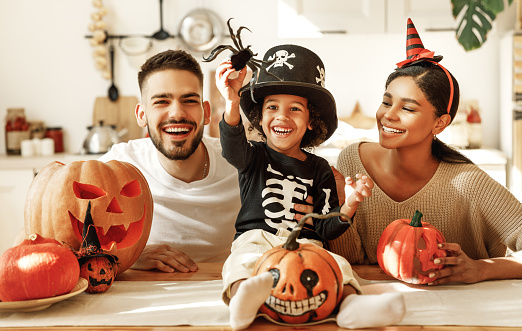A joyful family with a young child dressed in a pirate hat celebrating halloween. they are at a table with carved pumpkins and small decorative items, smiling and having fun together.
