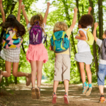 A group of six diverse children, viewed from behind, joyfully jumping in the air with their arms raised on a sunlit forest path, all wearing backpacks.