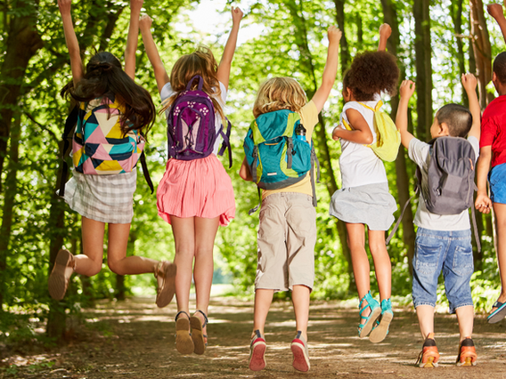 A group of six diverse children, viewed from behind, joyfully jumping in the air with their arms raised on a sunlit forest path, all wearing backpacks.
