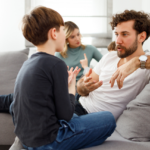 A father and son sit on a couch having a serious conversation, gesturing with their hands, while a woman looks on from the background.