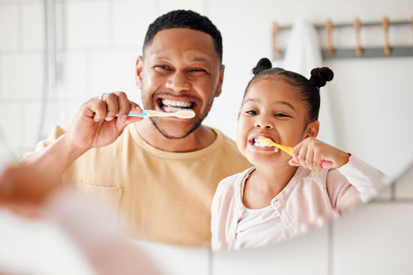 A joyful father and his young daughter brush their teeth together in a bright bathroom, both smiling broadly with toothbrushes in hand.