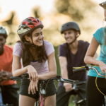 Four diverse friends enjoying a sunny day outdoors with their bicycles, smiling and conversing happily. one woman leans on her bike while chatting with another, both wearing helmets.