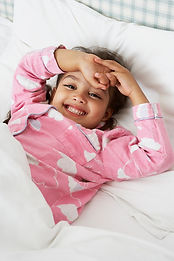 A cheerful young girl in a pink heart-patterned pajama top lies on a bed, arms raised with one hand on her forehead, smiling at the camera.