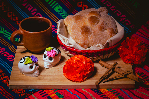A traditional day of the dead altar featuring a loaf of bread in a basket, two decorative sugar skulls, a clay cup, orange paper flowers, and cinnamon sticks, set against a colorful woven background.