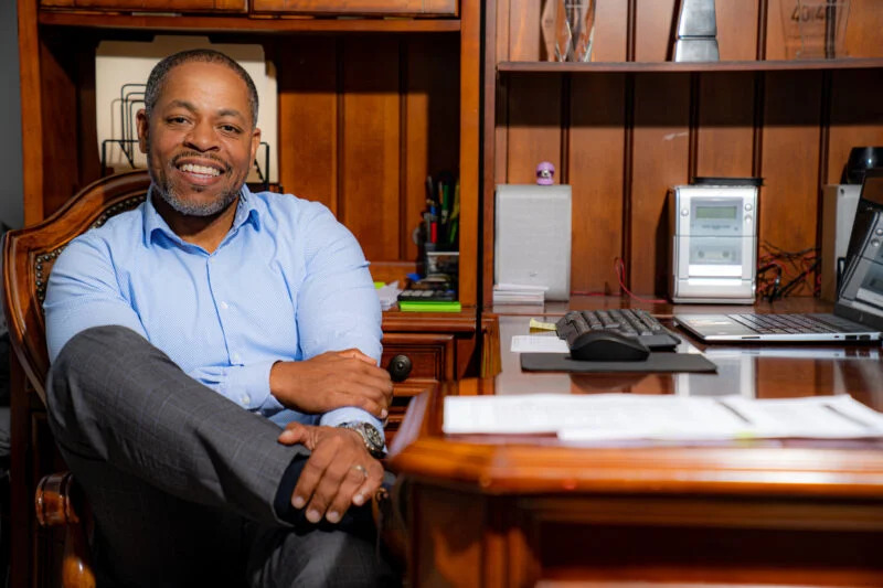 A man with a smiling expression, seated comfortably in a wooden office chair, wearing a blue shirt and jeans. he is in an office with shelves, a desk with papers, and a computer monitor.