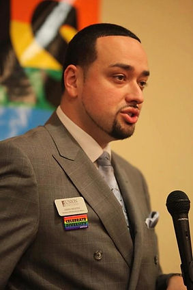 A man in a gray suit with a name tag speaks into a microphone, background featuring vibrant abstract art. he appears engaged and expressive.