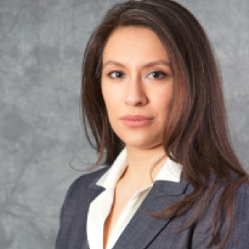 Professional portrait of a woman wearing a grey blazer over a white blouse, with shoulder-length brown hair, looking directly at the camera against a grey background.