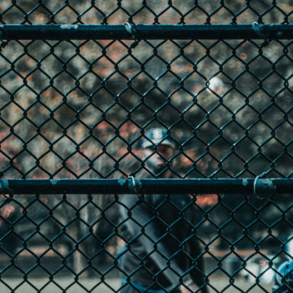 A person is partially obscured by a dark chain-link fence, with a blurred background showing an autumnal scene. the focal point is the fence's diamond pattern in silhouette.