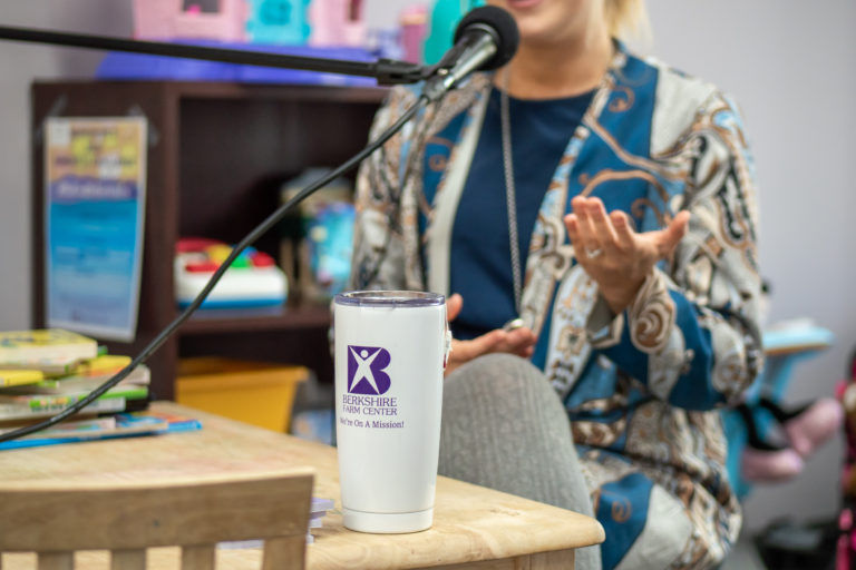 A woman speaking at a public event, gesturing with her hand, with a microphone and a promotional cup on the table in front of her. books are visible in the background.