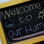 A small chalkboard with the words "welcome to our home" written in white, yellow, and pink chalk, decorated with a chalk-drawn cloud and heart.