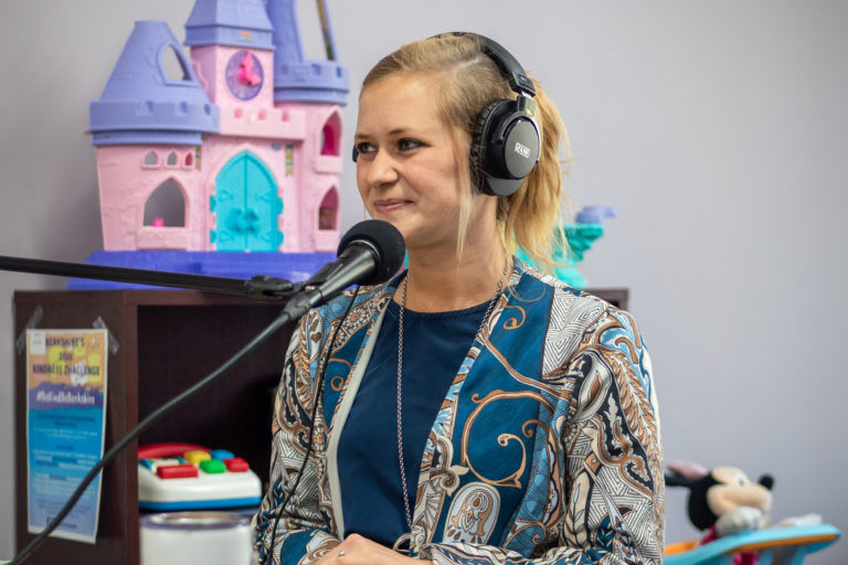 A woman in a patterned blouse wears headphones and speaks into a microphone, with a colorful toy castle and other toys blurred in the background.