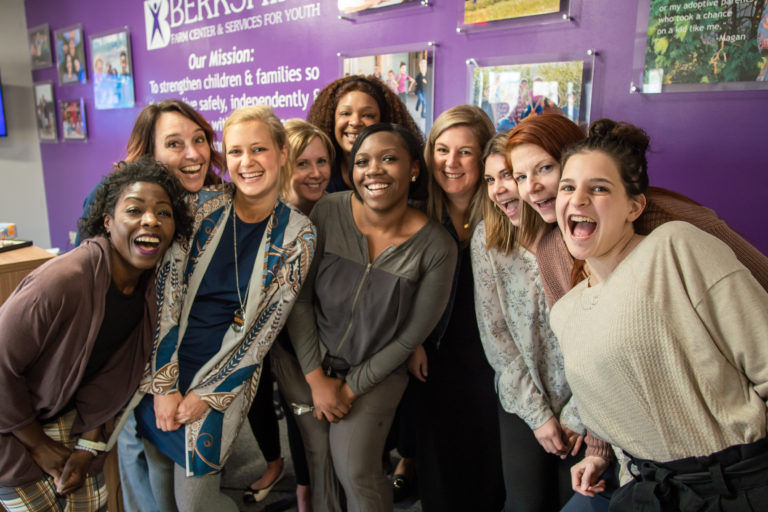 A group of nine diverse women smiling joyfully, standing together in a room with a purple wall. a poster with the text 