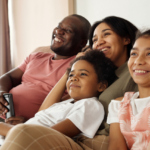 A happy family of four, with a father, mother, daughter, and son sitting closely together on a couch, smiling and watching tv in a brightly-lit living room.