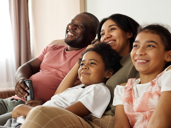 A happy family of four, with a father, mother, daughter, and son sitting closely together on a couch, smiling and watching tv in a brightly-lit living room.