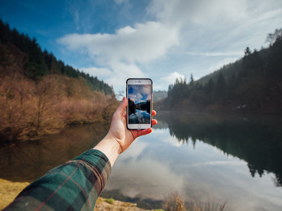 A person's hand holding a smartphone capturing a photo of a serene lake surrounded by forested hills under a partly cloudy sky.