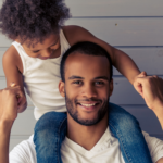 A joyful young father with a beard, wearing a white t-shirt and denim jacket, gives his small daughter a piggyback ride. she, in a white tank top, laughs while holding his hands for support.