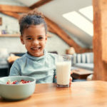 A smiling young girl with a ponytail sitting at a dining table, with a glass of milk in front of her and a bowl of cereal with fruits. the room has wooden beams and skylights.