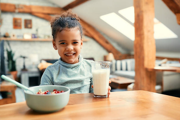 A smiling young girl with a ponytail sitting at a dining table, with a glass of milk in front of her and a bowl of cereal with fruits. the room has wooden beams and skylights.