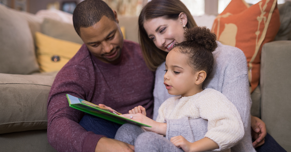 A young family enjoys reading together. a man and a woman sit on a couch with a toddler in between them, all focused on a colorful book the child is holding.