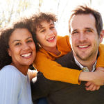 A joyful family outdoors with the father carrying the son on his back and the mother beside them, all smiling broadly. sunlight filters through trees in the background.