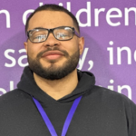 A man with glasses and a beard smiles gently, standing in front of a purple backdrop with text, wearing a black hoodie and a blue lanyard.
