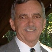 An older man with a warm smile, wearing a classic suit and tie, stands under a wooden shelter surrounded by lush foliage.