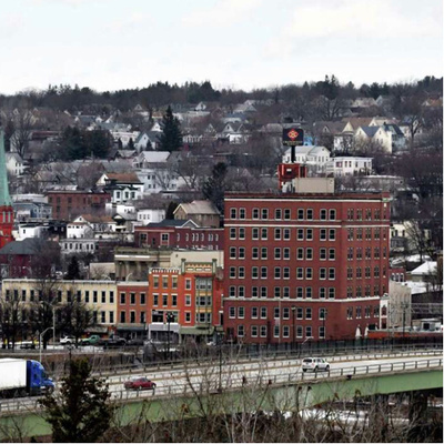 A multi-story red brick building stands prominently in a town with numerous houses and smaller buildings scattered in the background. A highway with vehicles passes in front of the building, and there is a mixture of bare trees and snow on the ground.