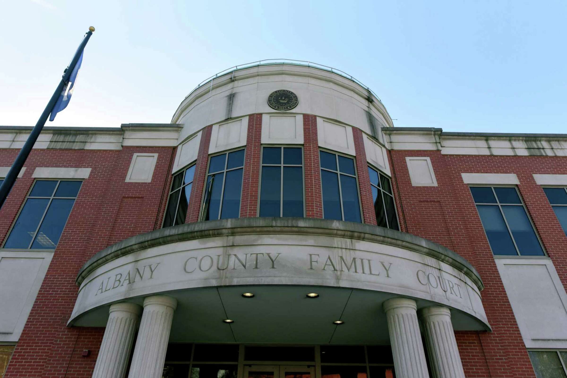 A front view of Albany County Family Court, a red-brick building with a circular entrance featuring white pillars and an inscription above that reads "ALBANY COUNTY FAMILY COURT". A flagpole with a flag is seen to the left of the entrance.