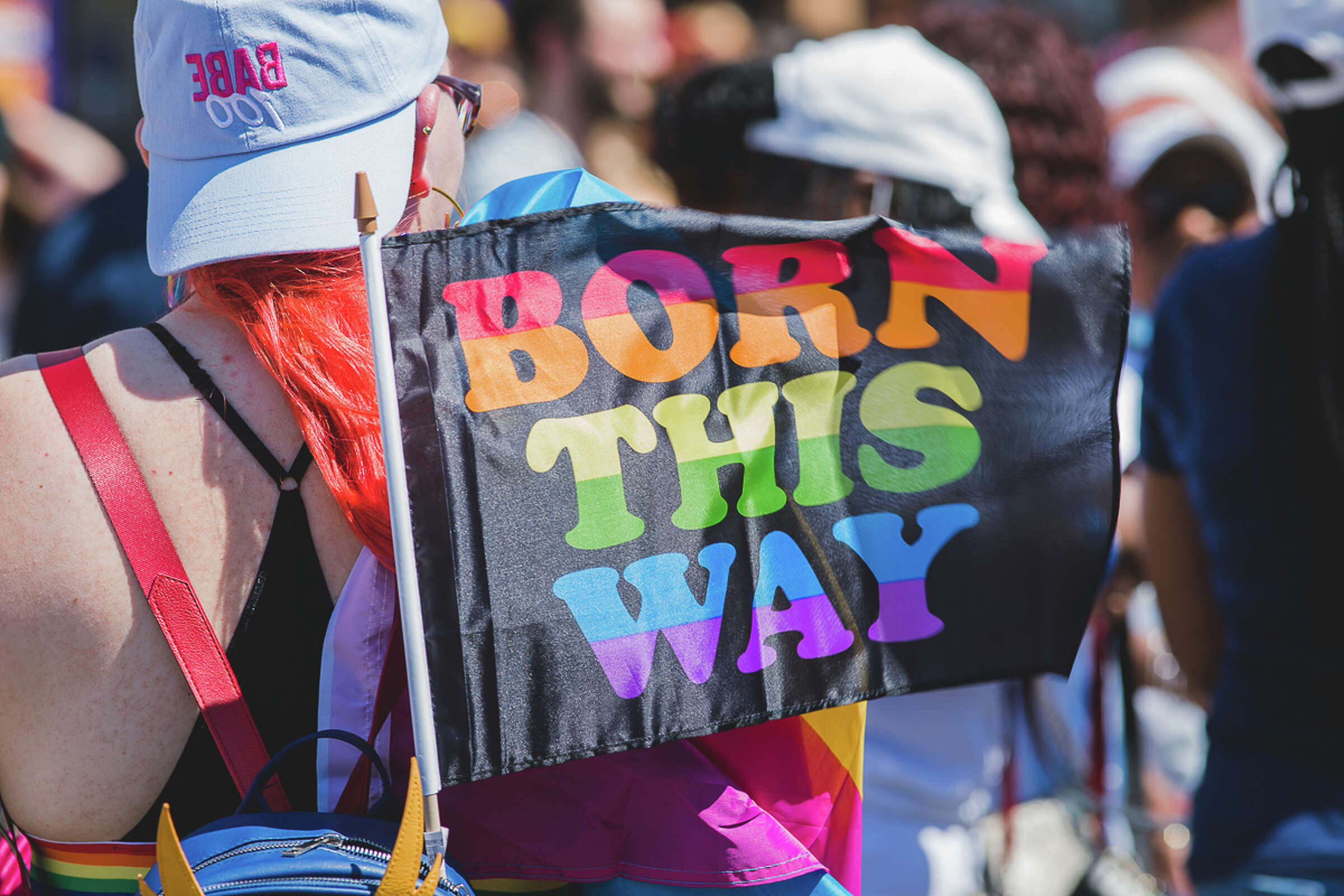 Person with bright red hair and a white cap at a pride event, holding a rainbow flag that reads "BORN THIS WAY." The scene is crowded with other attendees, some of whom are also wearing colorful attire.