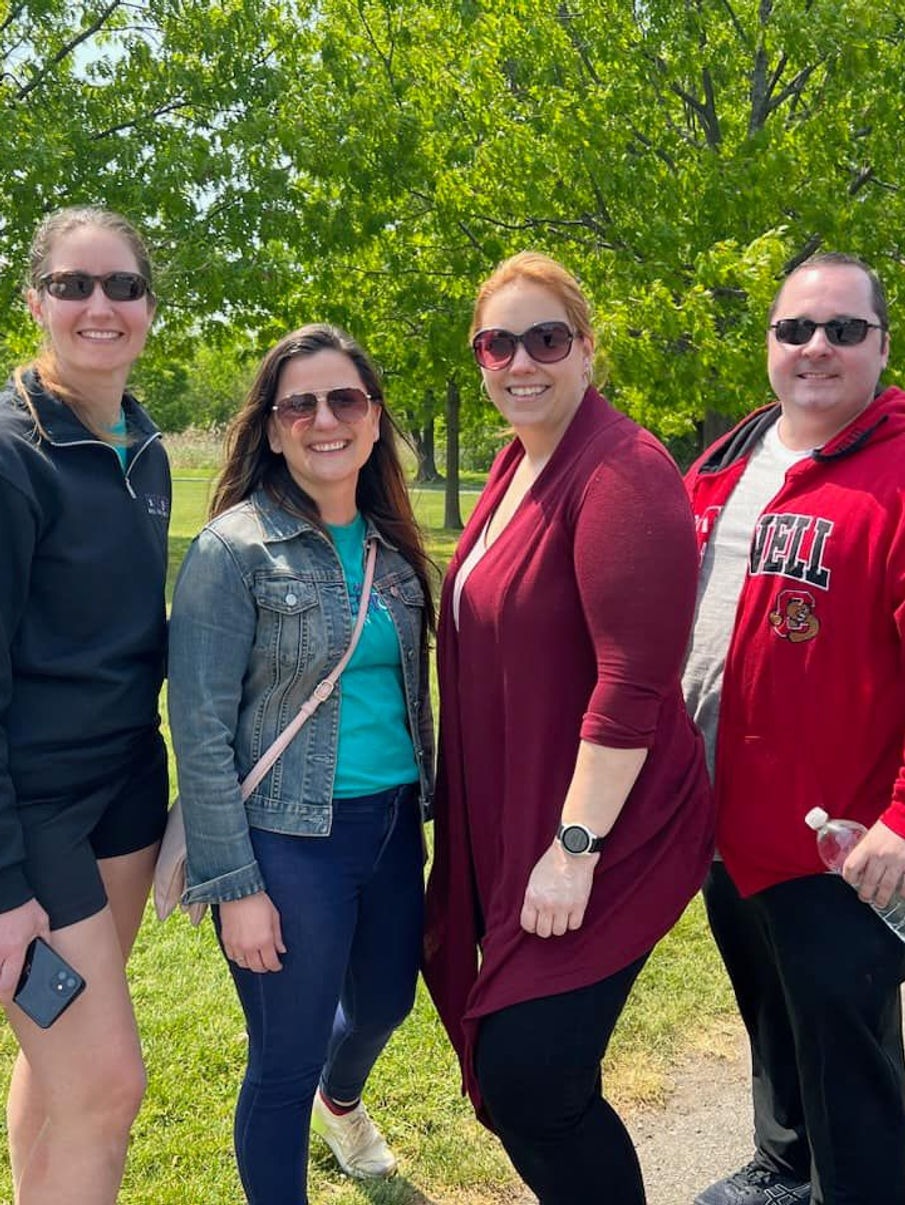 Four friends enjoying a sunny day in the park, three women and one man, smiling and wearing casual spring attire, with green trees in the background.