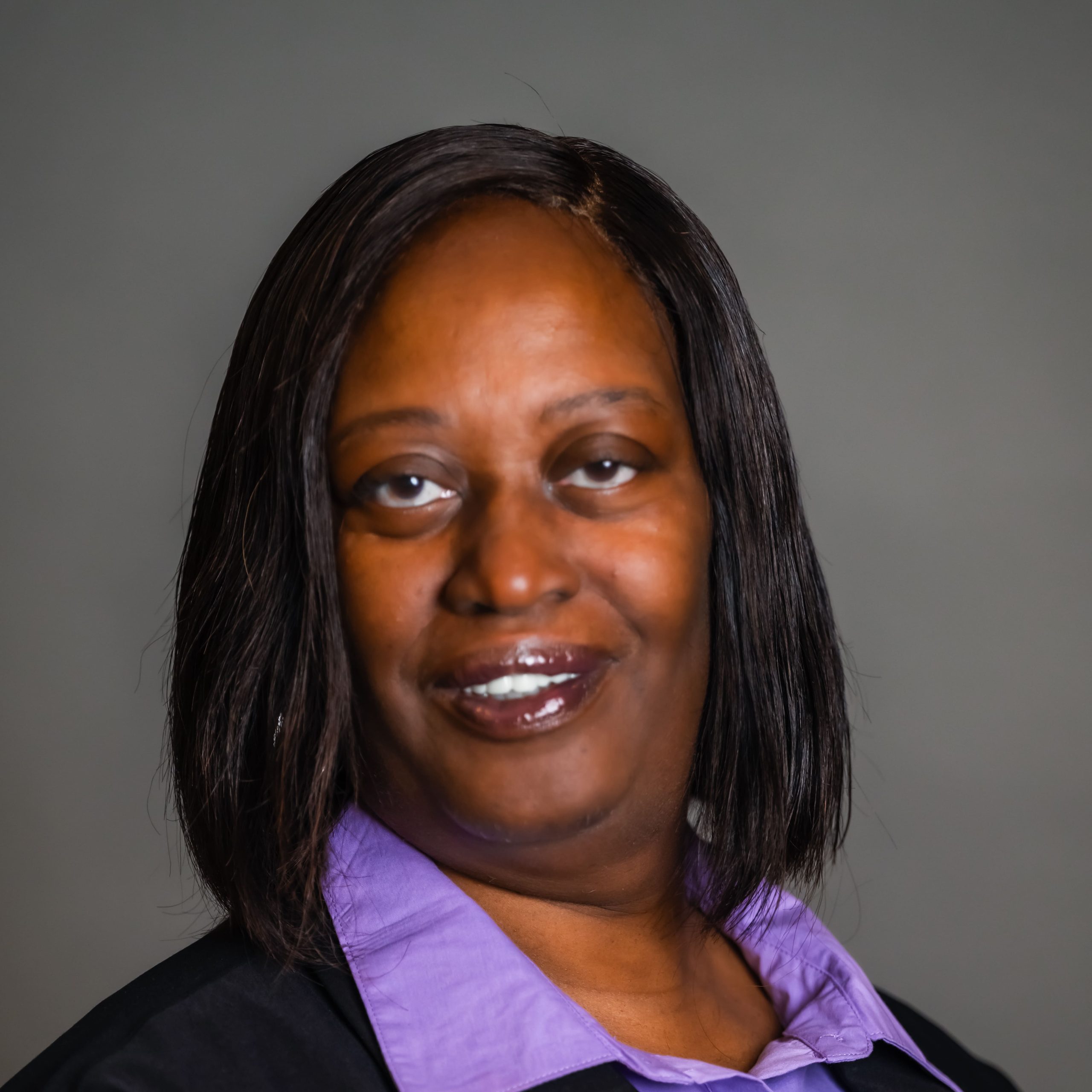 Jennifer with shoulder-length straight black hair smiles at the camera against a grey background. She is wearing a black blazer over a lavender collared shirt.