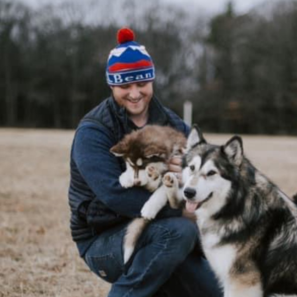 Matt kneels on the grass, smiling while holding a fluffy brown and white puppy. Next to them is a large, happy dog with a similar coat. Matt is wearing a red, white, and blue beanie, a dark jacket, and jeans. The background is a wooded area.