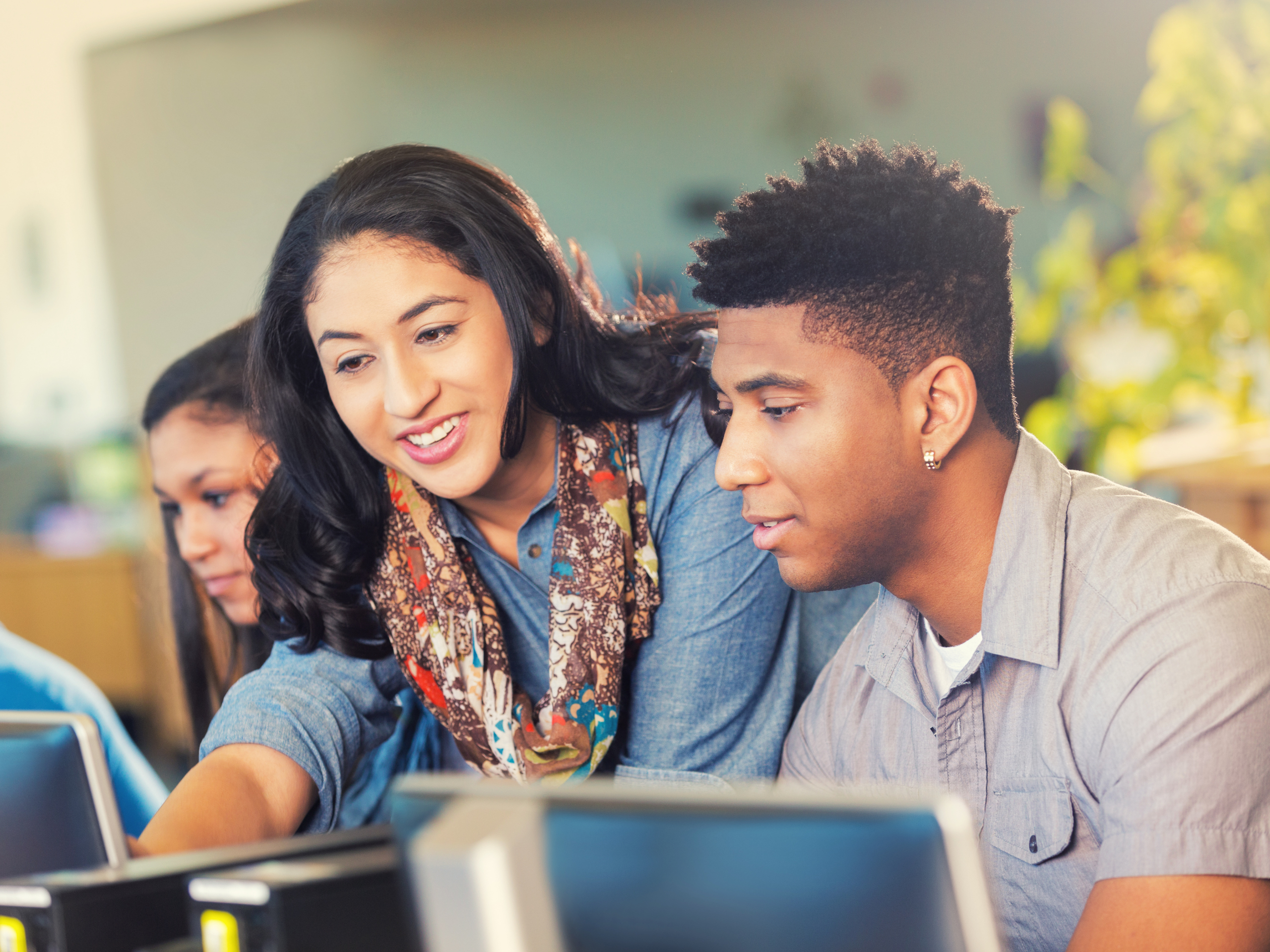 A group of young adults are seated at a desk, engaging with computers. One woman, smiling, is pointing at a screen while a man beside her attentively looks on. Another person in the background is also focused on a computer screen. They are in a well-lit room.