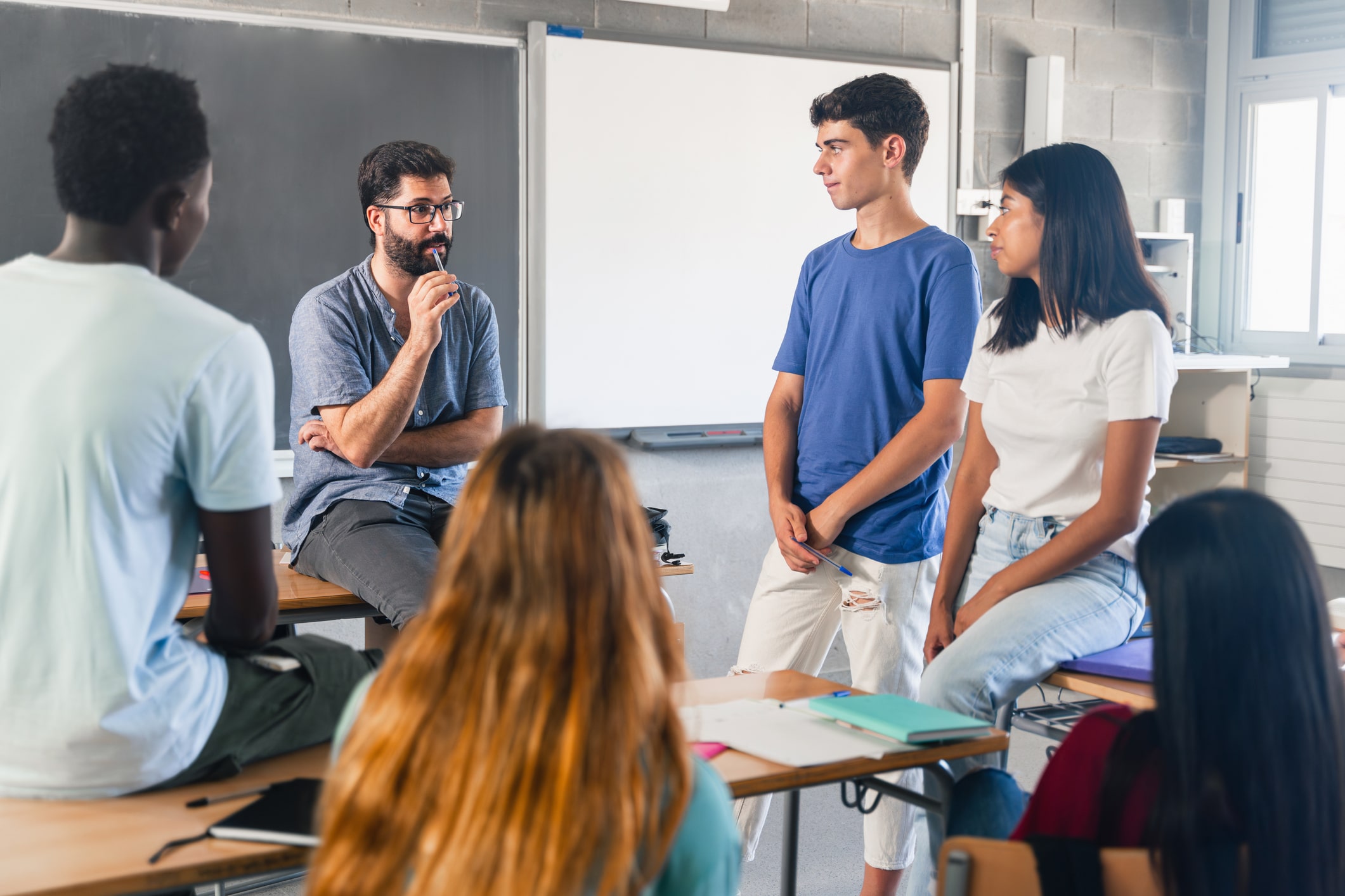 A classroom setting with a teacher sitting on a desk talking to a group of students. A whiteboard and a chalkboard are visible in the background.