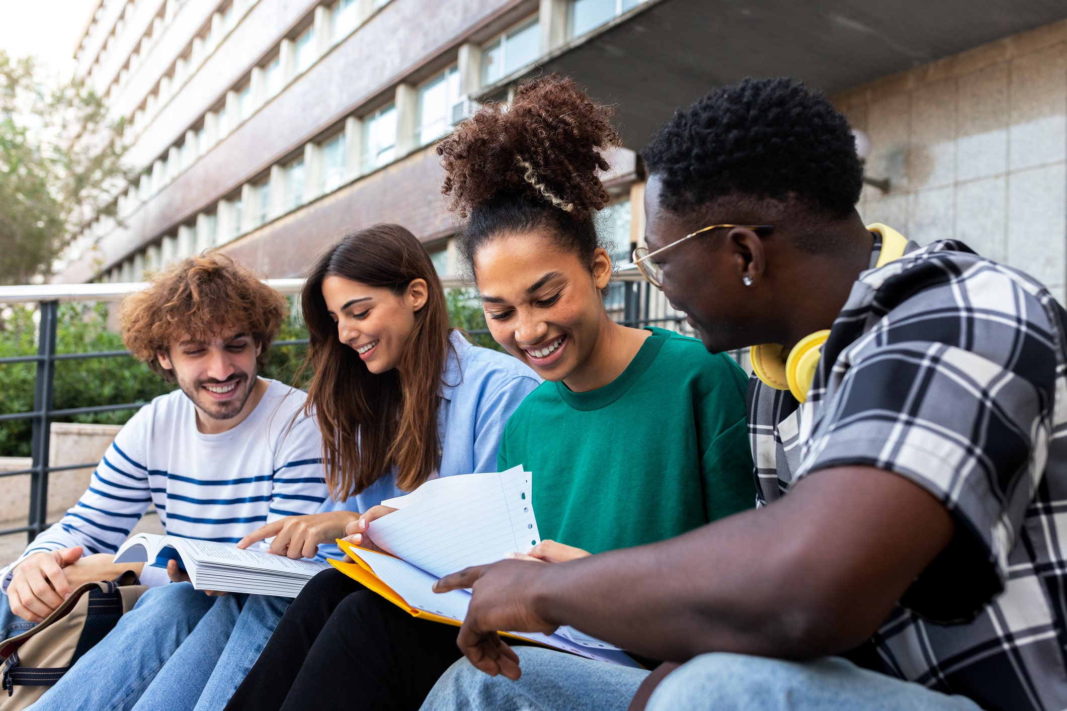 A group of four diverse young adults sit together outside on a sunny day. They are smiling and looking at notebooks and papers, possibly studying or sharing notes. Behind them is a multi-story building with large windows.