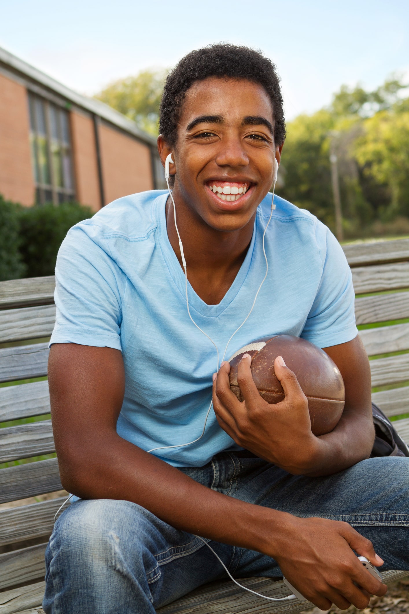 A young man sits on a wooden bench outdoors, wearing a light blue t-shirt and jeans. He is smiling, holding a football in one hand, and has white earphones in his ears. A brick building and trees are visible in the background.