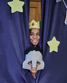 A joyful child wearing a crown peeks through a blue stage curtain decorated with paper stars, holding a paper theater mask in front of their face.