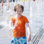 A young child wearing an orange shirt and blue tie-dye shorts is playing joyfully in a water fountain. They are smiling as water splashes around them on a sunny day. Other children and adults are visible in the background enjoying the fountain area.