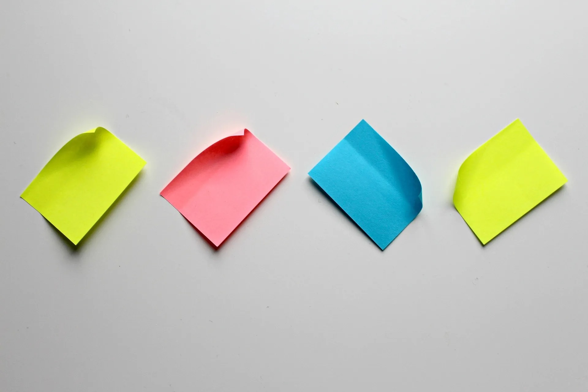 Four sticky notes, in neon yellow, pink, blue, and neon yellow, are arranged in a line on a white surface. Each sticky note has a corner slightly peeled, creating a casual, slightly three-dimensional effect.