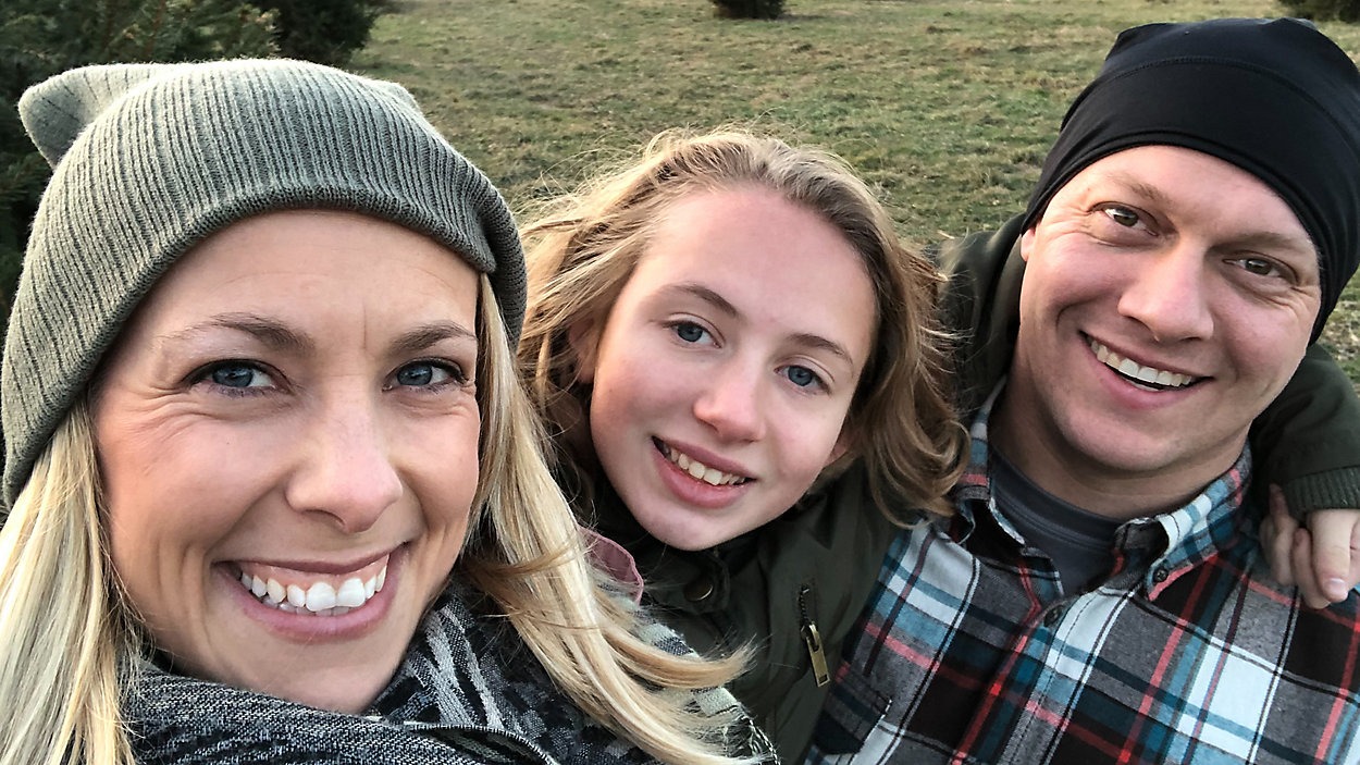 A group selfie features three people outside on a grassy field. They are dressed warmly, with the woman on the left wearing a gray knit beanie, the central person with long hair, and the man on the right in a black beanie and a plaid shirt. All smile at the camera.