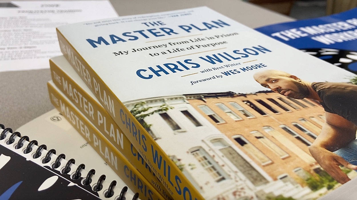 Close-up photo of a stack of "The Master Plan" books by Chris Wilson on a table, accompanied by a spiral-bound notebook. The cover features an image of Chris Wilson with a backdrop of row houses, and the subtitle reads "My Journey from Life in Prison to a Life of Purpose.