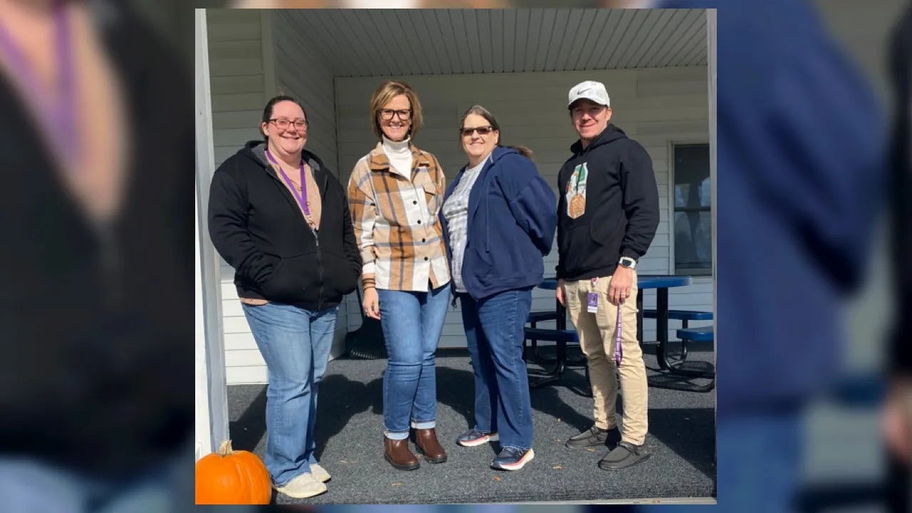 Four individuals stand together smiling outdoors in front of a building. They are casually dressed, with two wearing jackets and the others in sweaters. A small pumpkin is visible on the ground beside them, suggesting a fall scene.